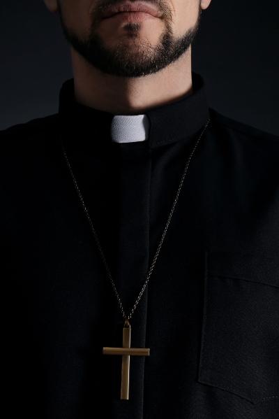 Close-up of a priest wearing a clerical collar and cross necklace against a dark background